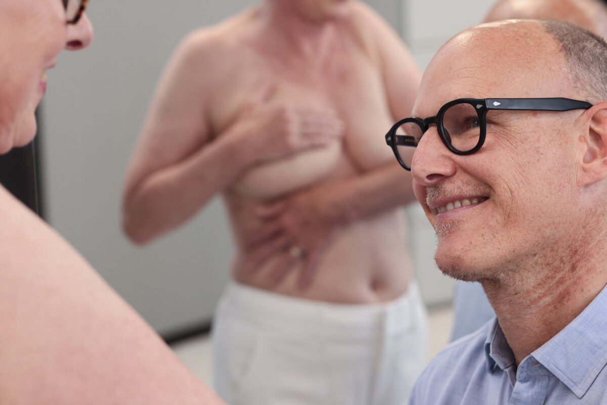 Frau wird vor einer Bruststraffung, Brustvergrößerung oder -verkleinerung bei Plastmed Düsseldorf untersucht von Dr. med. Till Scholz, der ihre Brust begutachtet und freundlich lächelt.